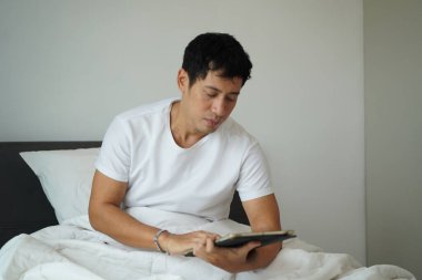 Asyalı adam yatakta oturup dijital tableti karıştırıyor. Sabah rutini, rahat ve odaklanmış ruh hali..
