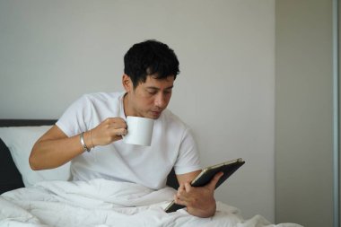 Yorgun ama odaklanmış Asyalı bir adam kahve içiyor ve sabahları yatakta dijital tablet kullanıyor..
