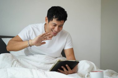 Dijital tablette video görüşmesinde yatakta gülümseyen adam el sallıyor. Sabah bağlantısı ve olumlu duygular.