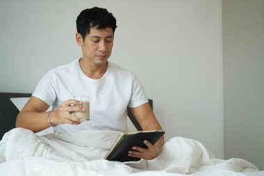 Asyalı adam beyaz tişört giyerek yatakta oturuyor, soya sütü içiyor ve tablet kullanıyor, evde sağlıklı yaşam tarzı gösteriyor.