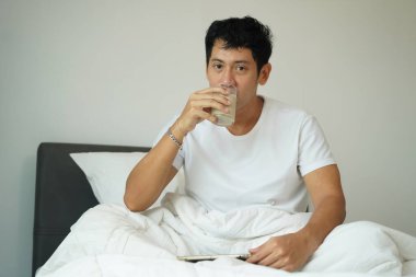 Beyaz gömlek giyen Asyalı adam soya sütü içiyor, yatakta dinlenirken kameraya bakıyor, huzurlu ve sağlıklı bir sabah.