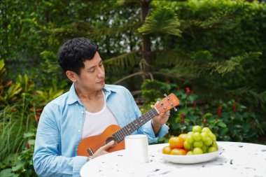 Asyalı bir adam bahçede beyaz masada oturmuş meyve tabağının yanında ukulele çalıyor ve huzurlu ve sağlıklı bir anın tadını çıkarıyor.