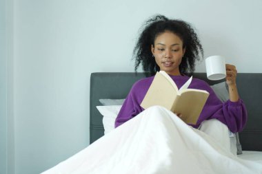 Genç Afrikalı kadın yatakta dinleniyor, elinde beyaz kupayla kitap okuyor, yumuşak doğal ışıkla rahat bir atmosfer.