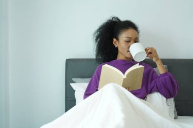 Genç siyah kadın kahvesini yudumluyor ve yatakta kitap okuyor, mor kazak giymiş, sessiz bir anın tadını çıkarıyor.