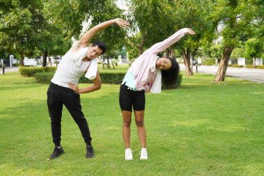 Asyalı erkek ve Afrikalı kadın, yeşil bir parkta yan yana esniyorlar, antrenmandan sonra gülümsüyorlar, sağlıklı yaşam tarzını ve vücut esnekliğini destekliyorlar.