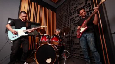 Üç Asyalı erkek müzisyen ses geçirmez müzik stüdyosunda elektronik gitar, bas ve davul çalıyor, modern iç mekanlarda canlı performans ve takım çalışmasına odaklanıyor