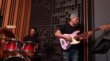 Asyalı erkek grup üyeleri ses geçirmez stüdyoda prova yapıyorlar, gitar, bas ve davul çalıyorlar ve kamera arkası sırasında doğal etkileşimle odak değiştiriyorlar.