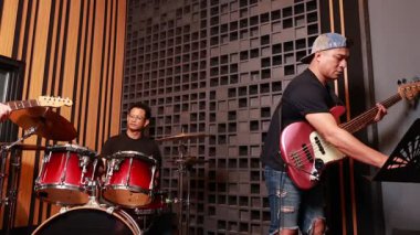 Asyalı erkek müzisyenler ses geçirmez kayıt stüdyosunda bas, davul ve gitar çalarken gülümsüyor ve ilgileniyorlar. Prova sırasında takım çalışmalarını ve eğlenmelerini gösteriyorlar.