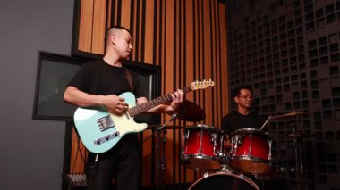 Asyalı erkek gitarist ve davulcu ses geçirmez stüdyoda birlikte çalıyorlar, senkronize enstrümanlar çalıyorlar, akustik ortamda günlük siyah kıyafetler giyiyorlar.