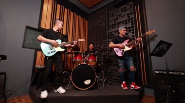 Üç Asyalı erkek müzisyen akustik stüdyoda konser veriyor, elektro gitar, bas ve davul çalıyorlar, gündelik modern kıyafetler giyiyorlar.