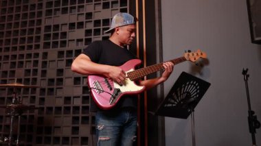 Asyalı erkek müzisyen ses geçirmez stüdyoda elektro bas gitar çalmaya odaklanmış, müzik standının yanında duruyor, iç mekanda günlük tarz kıyafetler giyiyor.