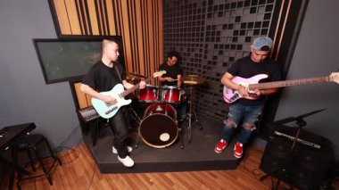 Profesyonel stüdyoda prova yapan Asyalı erkek müzisyenler, gitarist, davulcu, seans sırasında telefona basçı, müzik teçhizatı ile çevrili.