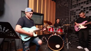Üç Asyalı genç müzisyen elektro gitar, bas ve davul çalarken konsantre oluyor. Kayıt stüdyosunda, ahşap akustik duvar, ses geçirmez arka plan, günlük giysiler.