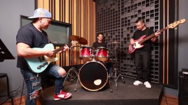 Profesyonel kayıt stüdyosunda elektronik gitar, bas ve davul çalan üç genç Asyalı erkek müzisyen, gülümsüyor ve performansın tadını çıkarıyor, ahşap akustik duvar, günlük kıyafet