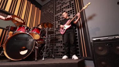 Üç Asyalı erkek müzisyen elektro gitar, bas ve davul setiyle kayıt stüdyosunda sahne alıyor. Çekim sahnesi, canlı atmosfer, günlük giyim, yaratıcı ses oturumu.