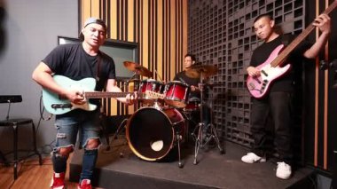 Profesyonel kayıt stüdyosunda canlı müzik yapan üç Asyalı genç adam, elektro gitar, bas ve davul çalıyor, odaklanmış ve ciddi ifadeler, ses geçirmez duvar