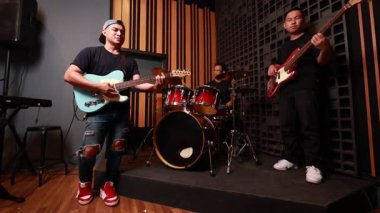 Üç Asyalı genç adam elektro gitarda, bas gitarda ve davulda akustik stüdyoda, ahşap ses geçirmez duvar, canlı ayakkabılar, günlük kıyafetler, neşeli ifade