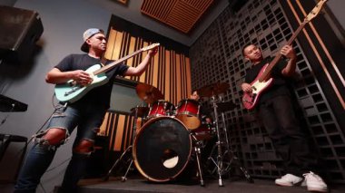 Üç Asyalı genç adam ses geçirmez stüdyoda elektro gitar, bas ve davul çalıyor. Odaklanmış ve tutkulu, gündelik kıyafetler, modern ekipmanlar, sıcak ışıklandırma.