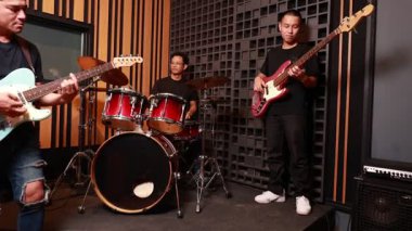 Profesyonel ses geçirmez odada sahne alan üç Asyalı erkek müzisyen, elektro gitarist, davulcu ve basçı prova sırasında birlikte çalan günlük kıyafetler giyiyorlar.