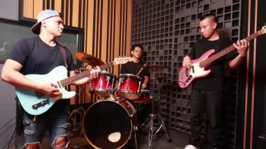 Profesyonel kayıt stüdyosunda müzik yapan üç Asyalı adam, enerjik çalışma oturumu, odak ve takım çalışması sırasında elektro gitar, pembe bas ve kırmızı davul seti çalıyorlar.