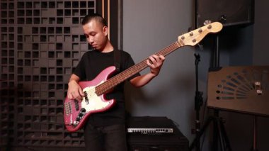 Profesyonel prova odasında metalik pembe bas gitar çalan genç Asyalı adam, odaklanmış ifade, ses ekipmanları ve müzik tezgahlarıyla çevrili.
