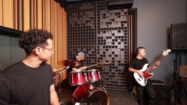 Modern müzik stüdyosunda prova yapan üç Asyalı adam, bas gitarist, davulcu ve grup lideri ses geçirmez arka planda odaklanma ve enerji ile işbirliği yapıyorlar.