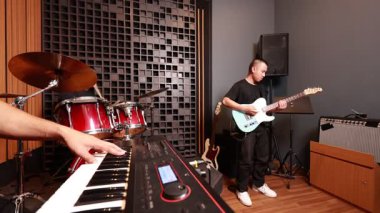 Asyalı klavyeci dijital tuşları çalarken, gitarist arka planda, bateri ve ses ekipmanları ile çevrili, dalgın reçel sahnesi