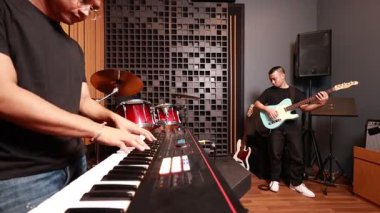 Asyalı bir erkeğin dijital klavye çalması, arka planda gitarist, kırmızı davul seti ve stüdyo ekipmanları görünür, müzikal sinerji