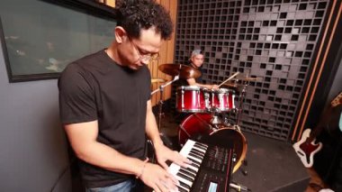 Asyalı erkek klavyeci bateristin önünde akustik prova stüdyosunun içinde gitar yansıması pencereden görülüyor, yaratıcı bir ortam.