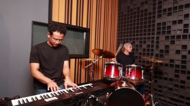 Asyalı klavyeci ve davulcu prova odasında ahşap ve akustik köpük arka planda müzik çalıyorlar. Rahat ve odaklanmış bir an.