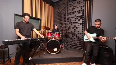 Akustik müzik stüdyosunda klavye, davul ve gitar çalan üç Asyalı erkek müzisyenin geniş açı, odaklanmış takım çalışması ve enerji