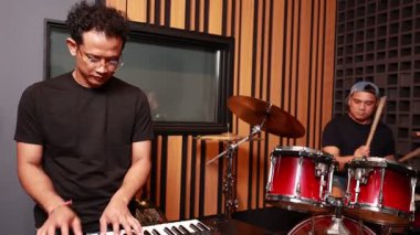 Üç Asyalı müzisyen akustik kayıt stüdyosunda klavye, davul ve elektro gitarla canlı performans sergileyerek takım sinerjisi ve duygularını gösteriyor.