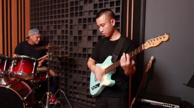 Asyalı gitarist ve davulcu ses geçirmez duvar ve ahşap panel arka planıyla profesyonel stüdyoda birlikte müzik çalıyorlar.