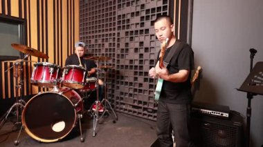 İki Asyalı erkek müzisyen, ses geçirmez bir müzik stüdyosunda elektro gitar ve kırmızı davul setiyle performans sergiliyorlar.