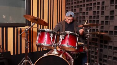 Genç Asyalı erkek davulcu gündelik kıyafetler giyip ses geçirmez stüdyoda kırmızı davul çalıyor, ritim ve müzik ifadesine odaklanıyor, arka plandaki stüdyo duvar panellerine odaklanıyor.