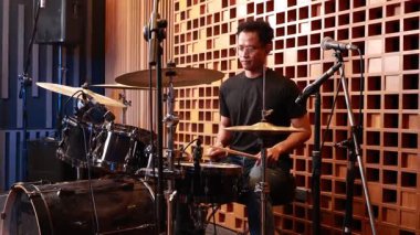 Genç Asyalı erkek davulcu ses geçirmez kayıt stüdyosunda davul çalıyordu. Mikrofonlar ve akustik panellerle çevriliydi ve müzik performansına odaklanmıştı.