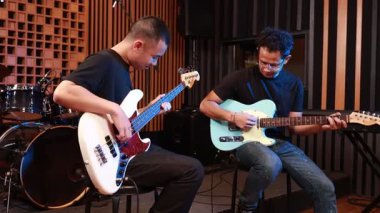 Modern kayıt stüdyosunda gitar ve bas çalan iki genç erkek müzisyen, biri kameraya gülümserken diğeri müzik çalmaya odaklanıyor, profesyonel müzik seansı.