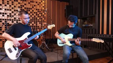 Profesyonel kayıt stüdyosunda bas ve elektro gitar çalan iki erkek müzisyen, arka planda akustik paneller, davul seti ve mikrofonlar ile konsantre ve yaratıcı ruh hali