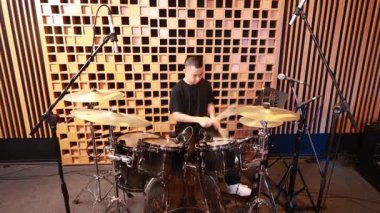 Genç Asyalı erkek davulcu, mikrofonlar ve ses geçirmez tahta paneller ile çevrili, akustik kayıt stüdyosunda davul seti üzerinde çalışırken odaklandı.