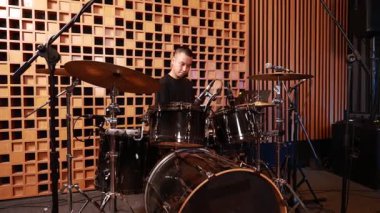 Çevresinde ahşap akustik paneller ve mikrofonlar olan profesyonel kayıt stüdyosunda siyah bateri arkasında oturan kendinden emin genç Asyalı erkek davulcu.