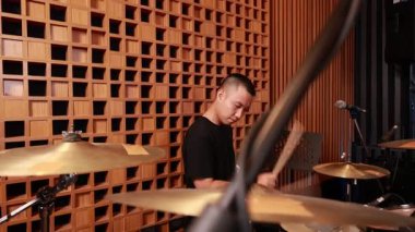Akustik kayıt stüdyosunda davul çalan genç Asyalı erkek davulcu bagetlerle zillere vuruyor, mikrofon ve ahşap panellerle kapalı alanda performans düzenliyor.