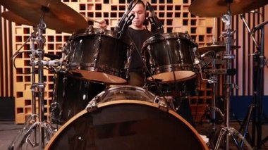 Asyalı genç adam ses geçirmez kayıt stüdyosunda davul çalıyor. Etrafı müzik ekipmanlarıyla çevrili, siyah tişörtlü, odaklanmış ve ciddi bir müzik prodüksiyonu ve performansı var.