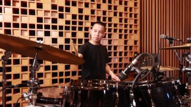 Siyah gömlekli odaklanmış Asyalı davulcu, mikrofonlar ve ahşap akustik paneller, dışavurumcu ve dinamik kapalı mekan performansı ile çevrili kayıt stüdyosundaki davul setinde enerjik ritim çalıyor.