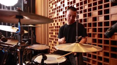 Genç Asyalı erkek davulcu profesyonel kayıt stüdyosunda, mikrofonlar ve akustik duvarlarla çevrili, canlı müzik sırasında odaklanmış ifade, kapalı sahne