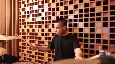 Profesyonel akustik kayıt stüdyosunda davul çalan Asyalı erkek davulcu. Canlı müzik sırasında odaklanmış ifade. Kapalı mekanda ışıklandırması olan ahşap duvar arka planı.