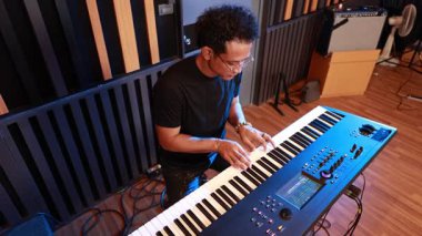 Profesyonel müzik stüdyosunda elektronik klavye çalan siyah tişörtlü genç Asyalı erkek müzisyen, görünür tuşlar, kontrol paneli ve el hareketleriyle performansa odaklanmış durumda.