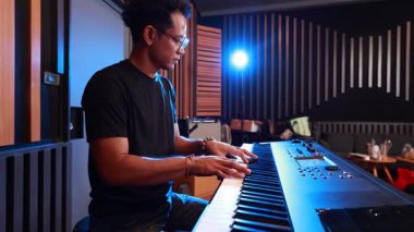 Modern kayıt stüdyosundaki dijital klavye ayarlarını ayarlayan siyah tişörtlü genç Asyalı adam, müzik yapmaya odaklanmış, arka plan ışığı ve yumuşak ruh haliyle kapalı çekim