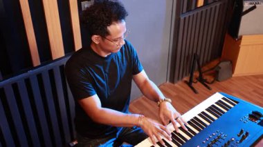 Asyalı bir adamın modern müzik stüdyosunda elektronik klavye çalarkenki resmi, odaklanmış ifade, görünür el tekniği ve ahşap paneller ve dişli akustik oda ortamı