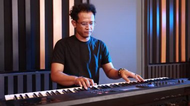 Yetişkin Asyalı adam ses geçirmez kayıt stüdyosunda çalarken dijital sentezleyicinin kontrol düğmelerini ayarlıyor, odaklanmış ifade, modern akustik panel arka planı