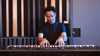 Asyalı müzisyen profesyonel kayıt stüdyosunda elektronik klavyede performans sergiliyor. Üzerinde siyah gömlek, odaklı ifade ve arka planda ses geçirmez duvar panelleri var.